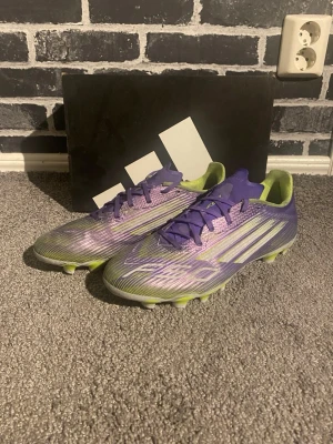 Lila Adidas X Speedflow fotbollsskor - Säljer ett par Adidas X Speedflow fotbollsskor i lila och neongult. Skorna har en strömlinjeformad design med snörning, syntetiskt material och mönstrad ovandel för bra bollkontroll. Sulan har fasta dobbar i neongult för optimalt grepp på gräsplan.
