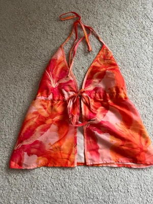 Blommig halterneck linnetopp - Snygg halterneck linnetopp med blommigt mönster i rött, orange och rosa. Toppen har knyt i nacken och framtill, samt en lätt utsvängd passform. Perfekt för varma sommardagar och festivaler.
