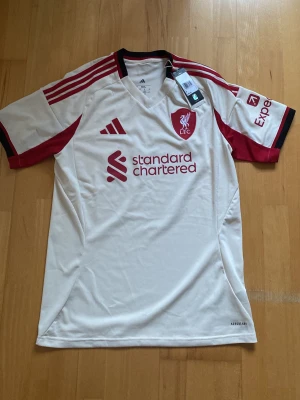 Liverpool FC bortatröja Adidas M - Snygg Liverpool FC bortatröja från Adidas i slim fit-modell. Tröjan är vit med röda detaljer, klubbmärke på bröstet och sponsorloggor. Tillverkad i polyester, perfekt för fotboll eller träning. Klassisk V-ringning och moderna ränder på axlarna.