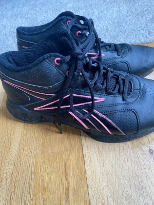 Svarta Reebok sneakers med rosa detaljer - Coola svarta sneakers från Reebok med rosa detaljer på sidorna och tungan. Skorna har klassisk snörning och är tillverkade i syntetmaterial med mesh på insidan för extra komfort. Perfekt för dig som vill ha en sportig och stilren look.