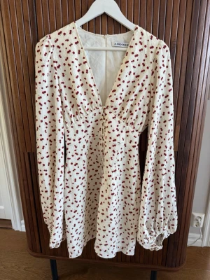 Vit blommig klänning från Adoore - Supersöt vit kortklänning ”Florence dress” från Adoore med röda blommor, v-ringning och långa puffärmar. Klänningen har en figurnära passform upptill och faller fint nedtill! OBS! Mönstret är som på de två bilderna där klänningen visss på galge. Bilden med klänningen ”på” är för att visa modellen ! Endast använd vid ett tillfälle. Nypris; 1495:-