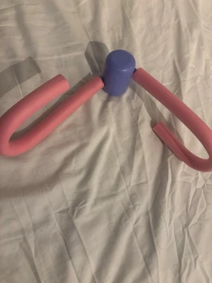 Träningsredskap lår- och armtränare - Ett smidigt träningsredskap i rosa och lila, perfekt för att träna lår, armar eller bröst. Tillverkad i skumgummi och plast, lätt att använda hemma eller ta med till gymmet. Enkel design med böjda handtag för bra grepp.
