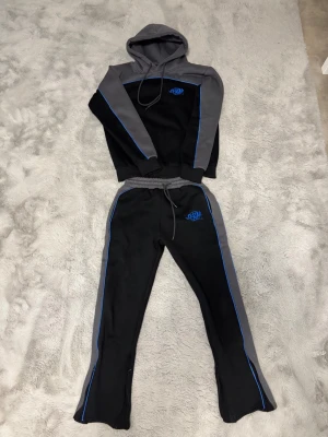 Syna World Tracksuit - Snyggt loungewear-set från Syna World i svart och grått med blå detaljer. Setet består av en hoodie med dragsko och matchande byxor med resår i midjan. Båda plaggen har blå piping och logga på bröst och höft. Perfekt för chill dagar hemma. TAGS FINNS