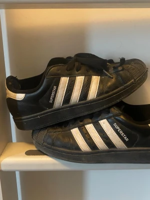 Adidas Superstar svarta sneakers - Klassiska Adidas Superstar sneakers i svart läder med vita detaljer och de ikoniska tre ränderna på sidan. Skorna har snörning, tåhätta i gummi och låg profil. Perfekta för dig som gillar en tidlös streetstil.
