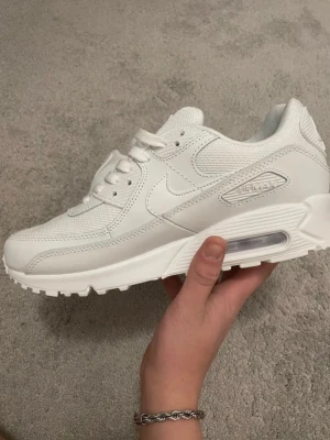Nike Air Max 90 vita sneakers - Snygga helvita Nike Air Max 90 sneakers med klassisk siluett. Skorna har ovandel i mesh och läder, synlig Air-enhet i sulan och ikoniska swoosh-loggan på sidan. Perfekta för dig som gillar stilrena och tidlösa sneakers.