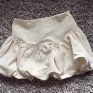 Krämfärgad ballongkjol från Gina Tricot - Superfin kort ballongkjol i krämfärgad satin från Gina Tricot i storlek 36. Kjolen har en hög midja och bubblig form som ger en trendig och lekfull look. Perfekt att styla med en enkel topp eller stickad tröja. Den är helt oanvänd med prislapp kvar 