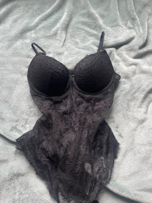 Svart spetsbody med tunna axelband - Snygg svart body i spets med vadderade kupor och tunna justerbara axelband. Bodyn har genomskinliga partier och en feminin, figurnära passform. Perfekt för dig som gillar en elegant och sexig stil.