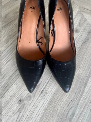 Svarta pumps med krokodilmönster H&M - Stilrena svarta pumps från H&M i storlek 40 med spetsig tå och krokodilmönstrad yta. Skorna har en klassisk klack och är tillverkade i konstläder med brun innersula. Perfekta för dig som gillar en trendig och elegant look.