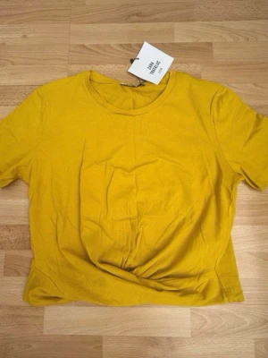 Ny. Gul t-shirt från Zara Trafaluc - En basic gul t-shirt med knut knyt fram från Zara Trafaluc med rund halsringning och korta ärmar. Tillverkad i mjuk bomull, perfekt för en avslappnad och trendig look. Enkel att matcha med jeans eller kjol.
