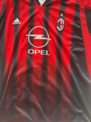AC Milan Adidas matchtröja röd/svart - Klassisk AC Milan matchtröja från Adidas i rött och svart med smala svarta ränder. Broderat klubbmärke, gul stjärna och Adidas-logga på bröstet. Opel som sponsor framtill. Tillverkad i lätt och ventilerande polyester, perfekt för fotbollsträning eller match.