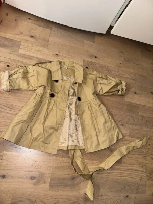 Beige trenchcoat med knytskärp - Stilren beige trenchcoat med dubbelknäppning och tillhörande knytskärp i midjan. Jackan har vida ärmar med uppvikta muddar och ett glansigt innerfoder. Perfekt för en trendig och klassisk look.Beige trenchcoat med knytskärp