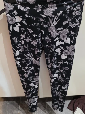 Svarta leggings med blommigt mönster - Snygga svarta leggings med vitt och grått blommönster över hela byxan. Stretchigt material som sitter tajt och bekvämt. Perfekta för träning eller chill dagar. Hög midja för extra komfort och stil.