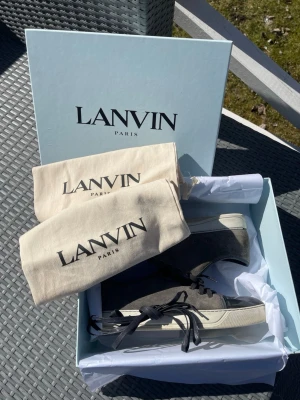 Lanvin grå låga sneakers - Snygga sneakers från Lanvin i grå mocka med svart tåparti i blankt läder och vita sulor. Klassisk låg modell med svarta skosnören och beige innersula. Kommer med originalkartong och dustbags. Perfekta för dig som gillar stilrena och exklusiva sneakers.