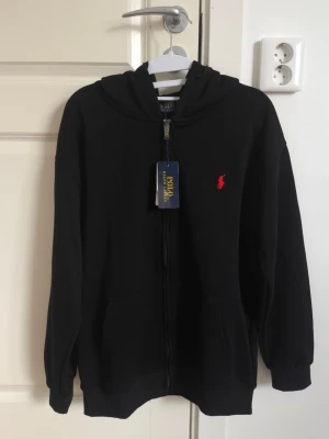 Polo Ralph Lauren Zip Hoodie - Svart - Helt ny med tags -     Säljer en stilren och klassisk tröja med dragkedja från Polo Ralph Lauren. Tröjan är i nyskick, aldrig använd — säljer eftersom storleken inte passar.    Den har en bekväm huva med två dragsnören samt fickor framtill i mitten, vilket gör den både praktisk och skön att använda.        Mått (ca): Axelbredd: 53 cm Längd: 65 cm.               Logotyp: Klassisk broderad logotyp i rött på bröstet.     Material: Mjuk och högkvalitativ bomullsblandning med fleece inuti.