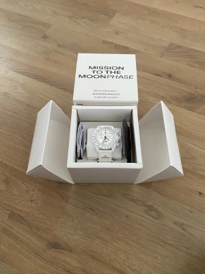 Omega x Swatch Moonswatch Mission To The Moonphase - Snygg Omega x Swatch MoonSwatch Mission to the Moonphase i helvitt biokeramiskt material. Klockan har vit urtavla med svarta detaljer, tachymeter och månfaskomplikation. Matchande vitt band med Omega-logga. Perfekt för dig som gillar stilrena accessoarer.