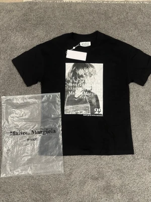 Maison Margiela svart t-shirt med print - Svart t-shirt från Maison Margiela med vit grafisk print framtill och klassisk rund hals. T-shirten har korta ärmar och är tillverkad i mjuk bomull. Perfekt för dig som gillar streetstyle och vill sticka ut med en unik design.