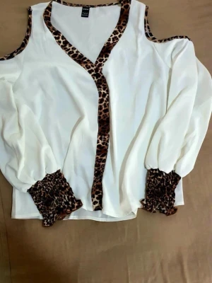 Vit blus med leoparddetaljer från shien - Snygg vit blus från SHEIN  med leopardmönstrade detaljer längs v-ringningen, axelbanden och vid de vida ärmsluten. Blusen har cold shoulder-design och är tillverkad i ett lätt, flytande material som ger en avslappnad men trendig look.