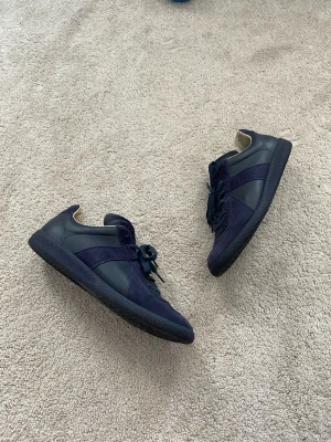 Marinblå sneakers Maison Margiela - Äkthetsverifierade maison margiela Gats. Snygga marinblå sneakers från Maison Margiela med ovandel i både mocka och läder. Klassisk låg modell med svarta snören och gummisula. Diskret logga under sulan och beige innerfoder för extra komfort.