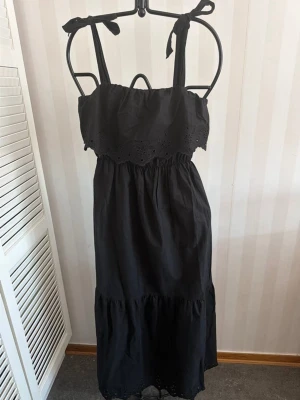 Svart långklänning med knytaxelband - Svart långklänning med broderade detaljer och volang upptill. Klänningen har knytbara axelband och resår i midjan för en bekväm passform. Tillverkad i bomull, perfekt för varma dagar.