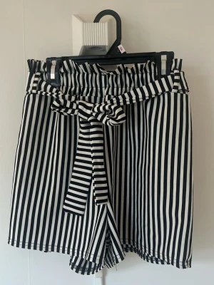 Randiga shorts med knytband - Snygga svartvita shorts med vertikala ränder och högt papperbag-midja. Knytband i midjan ger en extra detalj och shortsen har en lös och bekväm passform. Perfekta för varma dagar och en trendig look.