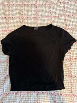 Svart ribbad topp från Gina Tricot - Svart ribbad topp från Gina Tricot med korta ärmar och en snygg cut out-detalj vid halsen. Toppen har en croppad passform och är perfekt för att lyfta vilken outfit som helst. Enkel att matcha med både jeans och kjol.