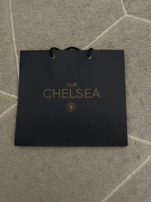 Chelsea Club shoppingväska i svart - Snygg shoppingväska från Chelsea FC i svart med guldfärgad text och logga. Väskan har rejäla svarta snören som handtag och är tillverkad i kraftigt papper. Perfekt för dig som gillar fotboll och vill bära dina grejer med stil.