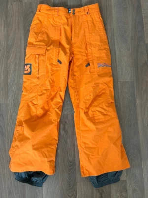 Orangea skidbyxor från Burton - Säljer ett par orangea skidbyxor från Burton med flera fickor, justerbar midja och förstärkta benslut. Finns några märken längst ner på benen samt något missfärgning på fickorna baktill. Storlek XL.