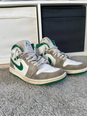 Air Jordan 1 Mid vit/grön/grå - Snygga Air Jordan 1 Mid sneakers i vitt läder med gråa mockadetaljer och gröna accenter på swoosh, häl och logga. Klassisk siluett med perforerad tå och gröna detaljer på sulan. Perfekta för dig som gillar streetwear och vill sticka ut. Ny pris 3000 kr. Det har blivit två knutar på ena skosnöret tyvärr men funkar ändå
