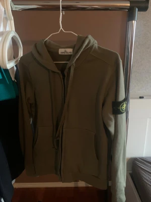 Olivgrön hoodie från Stone Island - Säljer en olivgrön hoodie från Stone Island med dragkedja framtill och klassisk patch på vänster ärm. Hoodien har fickor på sidorna och en skön huva. Perfekt för dig som gillar streetwear och vill ha något stilrent men ändå ikoniskt.
