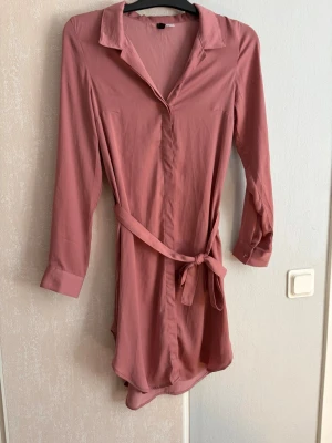 Rosa skjortklänning med bälte - Snygg rosa skjortklänning med lång ärm och knytbälte i midjan. Klänningen har klassisk krage och knappar framtill, perfekt för en trendig och avslappnad look. Materialet känns mjukt och följsamt mot kroppen.