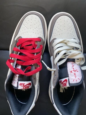 Nike Air Jordan 1 x Travis Scott Low - Säljer ett par Nike Air Jordan 1 Low Travis Scott med grå, vit och svart färgkombination. Skorna har en omvänd svart swoosh, röd och vit text på plösen samt olika färgade skosnören (röd och vit). Ovandelen är i läder och mocka för en lyxig känsla.