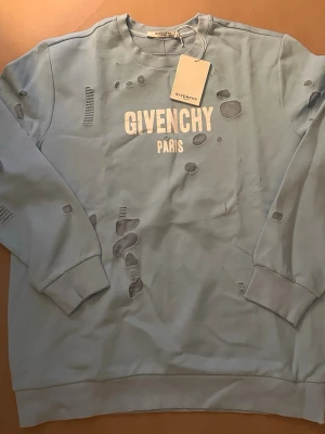 Ljusblå Givenchy Paris sweatshirt - Ljusblå sweatshirt från Givenchy Paris med distressed detaljer och tryckt logga framtill. Tröjan har rund halsringning, ribbade muddar och är tillverkad i mjukt bomullsmaterial. Perfekt för en trendig och avslappnad streetwear-look.