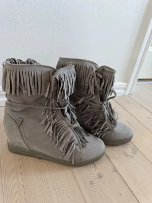 Gråa frannskor - Säljerrå boots med kilklack och massor av fransar framtill och upptill. Skorna är i mocka och har snörning framtill. Säljer även ett par i rosa. Hör av er om ni har frågor🩷