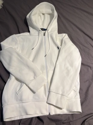 Vit hoodie från Polo Ralph Lauren - Snygg vit hoodie från Polo Ralph Lauren med dragkedja framtill och klassisk huva med dragsko. Broderad logga på bröstet och två fickor framtill. Tillverkad i mjukt bomullsmaterial som känns riktigt skönt mot huden. Perfekt för en clean och stilren look. Jätte fint skick, kan diskutera pris vid snabb affär 