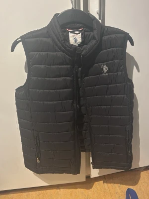 Svart dunväst från U.S. Polo Assn. - Stilren svart dunväst från U.S. Polo Assn. med quiltad design och hög krage. Västen har dragkedja framtill, två fickor med dragkedja och en broderad logga på bröstet. Perfekt för lager-på-lager och enkel att matcha med olika outfits.