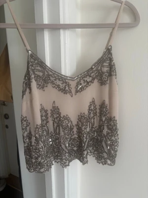 Beige paljettprydd linnetopp - Säljer en beige linnetopp med tunna axelband och vacker paljettbrodyr i silver. Toppen har en croppad passform och detaljerade mönster längs nederkant och byst. Perfekt för dig som vill sticka ut med en unik och glittrig topp.