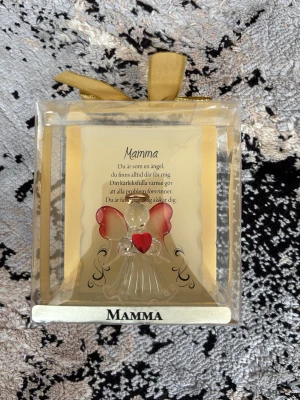 Glasängel med text till mamma - Handblåst glasängel med röda vingar och hjärta, levereras i en genomskinlig ask med en fin text till mamma. Perfekt present eller dekoration. Ängeln står på en platta med texten 'Mamma'.