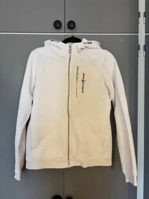 Vit hoodie från Sail Racing - Snygg vit hoodie från Sail Racing med dragkedja framtill och en bröstficka med logga. Hoodien har huva och två sidofickor. Tillverkad i mjukt material som känns skönt mot huden. Perfekt för dig som gillar stilren och sportig look.