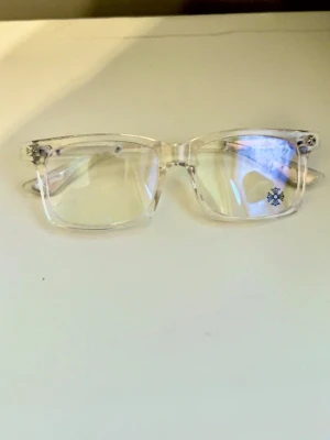 Chrome hearts glasögon silver med blått tonat glas  - Snygga chromehearts glasögon med transparent båge och rektangulära linser. Sidorna har silvriga detaljer och dekorativa mönster, vilket ger en unik och trendig look. Perfekt accessoar för att lyfta din stil.