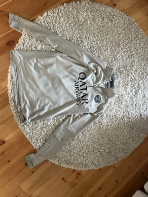 Beige PSG träningströja från Puma - Snygg beige långärmad träningströja från PSG med svart Qatar Airways-tryck på bröstet och PSG-logga. Tröjan har halv dragkedja vid halsen och svart signatur på ena ärmen. Perfekt för fotbollsträning eller sportiga dagar.
