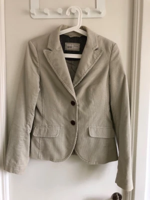 Beige kavaj i manchester från Esprit - Stilren beige kavaj i manchestertyg från Esprit med två knappar framtill och klassisk krage. Kavajen har två fickor med lock och är helfodrad med snyggt mönstrat innerfoder. Perfekt för en trendig och avslappnad look.
