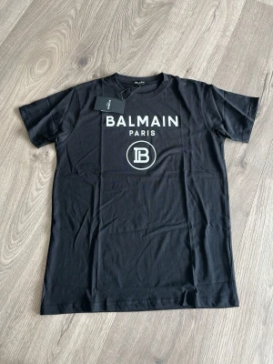 Svart Balmain Paris t-shirt - Snygg svart t-shirt från Balmain Paris med vit logga och tryck framtill. Klassisk passform med rund hals och korta ärmar. Perfekt för dig som vill ha en stilren och trendig look.