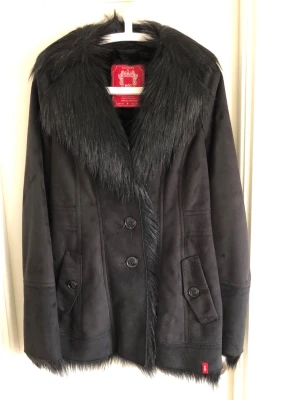 Svart kappa med fuskpäls från Esprit  - Stilren svart kappa från Esprit med bred krage och detaljer i svart fuskpäls. Kappan har knäppning med stora knappar framtill och två fickor med lock. Materialet är mjukt och matt, vilket ger en lyxig känsla. Perfekt för kalla dagar.