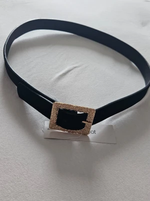 Svart bälte med glittrigt spänne - Stilrent svart bälte med ett fyrkantigt, glittrigt spänne i guld. Perfekt accessoar för att lyfta din outfit med en touch av glamour. Bältet är smalt och enkelt att matcha med olika looks. Ny med prislapp.