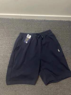 Marinblå shorts från Polo Ralph Lauren - Snygga marinblå shorts från Polo Ralph Lauren med klassisk logga broderad på benet. Shortsen har elastisk midja med snörning för skön passform. Perfekta för sommaren och tillverkade i mjukt material.