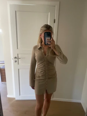 Beige/Guld kort skjortklänning med knappar - Trendig beige/guld skjortklänning med långa ärmar och knappar hela vägen framtill. Klänningen har en figurnära passform och snygg rynkdetalj längs mitten. Perfekt för dig som gillar stilrena och moderna plagg. Storlek S från bikbok!💞