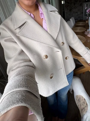 Kort cremefärgad ullrock - Snygg kort dubbelknäppt rock i cremefärgad ullmix med breda slag och dekorativa knappar framtill. Perfekt för dig som vill ha en stilren och trendig look. Passar till jeans eller kostymbyxor.