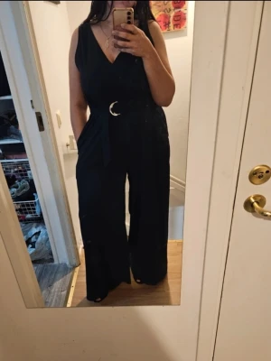 Mörkblå  ärmlös jumpsuit med bälte - Snygg svart ärmlös jumpsuit med v-ringning och vida ben.    Använd endast en gång så den ör som NY. I storlek M och passar storlek L då den är lite översize. Materialet ser ut att vara mjukt och följsamt.