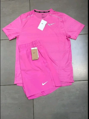 Nike rosa träningsset t-shirt & shorts - Nike träningsset i stark rosa färg med matchande t-shirt och shorts. T-shirten har rund hals, korta ärmar och diskreta ränder i materialet. Shortsen har elastisk midja och båda plaggen har vit Nike-logga. Tillverkade i lätt, snabbtorkande syntetmaterial.