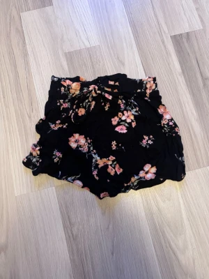 Svarta blommiga shorts med volang - Svarta shorts med blommigt mönster i rosa och orange toner. De har en lös passform, volangdetaljer och en knytning i midjan. Perfekta för varma dagar och ger en avslappnad men trendig look.